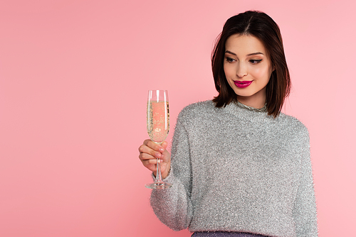 Brunette woman in sweater looking at glass of champagne isolated on pink