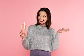 Smiling woman in sweater holding glass with champagne and pointing with hand isolated on pink
