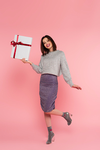 Positive woman in skirt and sweater holding present on pink background