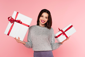 Happy woman in sweater holding presents and looking at camera isolated on pink