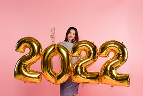Smiling woman holding glass of champagne near balloons in shape of 2022 isolated on pink