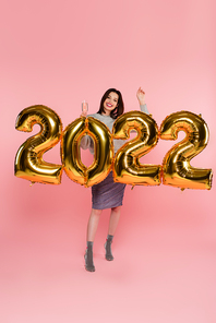 Pretty and stylish woman holding champagne near balloons in shape of 2022 on pink background