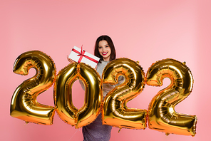 Smiling woman holding gift box near balloons in shape of 2022 numbers isolated on pink
