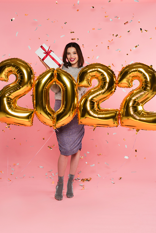Smiling woman holding present near balloons in shape of 2022 and confetti on pink background