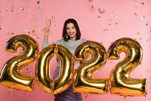 Cheerful woman holding champagne near balloons in shape of 2022 and confetti on pink background