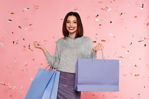 Stylish woman in sweater holding shopping bags under confetti on pink background