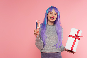 Stylish woman with dyed hair holding champagne and gift box isolated on pink