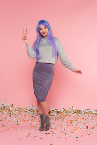 Happy stylish woman with dyed hair holding champagne near confetti on pink background