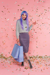 Positive woman with dyed hair holding shopping bags and waving hand under confetti on pink background
