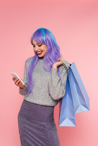 Happy woman with dyed hair using smartphone and holding shopping bags isolated on pink