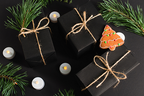 top view of gingerbread cookie on wrapped dark gift boxes near candles and pine branches on black