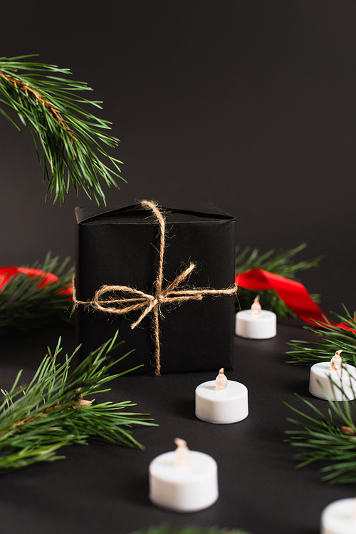 wrapped gift box near fir branches and candles with blurred red ribbon on black background