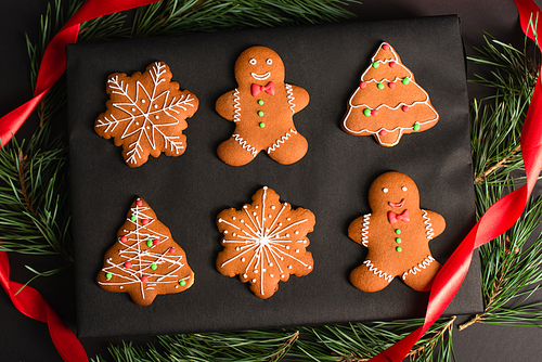 top view of different shapes gingerbread cookies on box near pine branches and red ribbon on black
