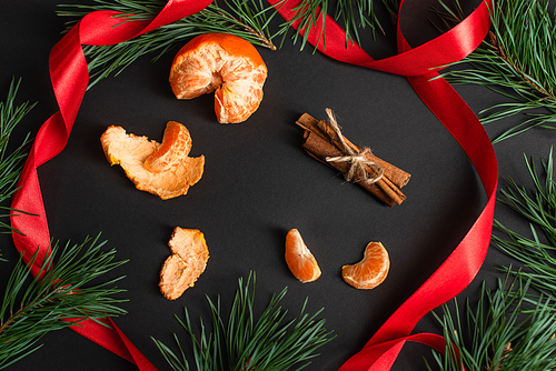 top view of peeled tangerines and cinnamon sticks near red ribbon and fir branches on black