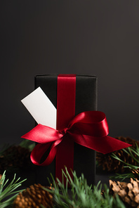 wrapped gift box with red ribbon and blank card near blurred fir branches and pine cones on black
