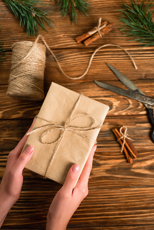cropped view of woman holding wrapped gift box near scissors on wooden surface