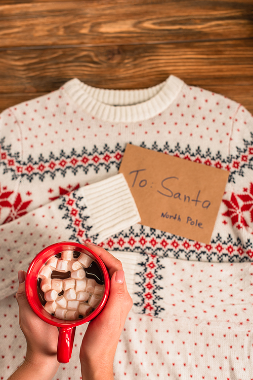 cropped view of woman holding cocoa with marshmallows near blurred letter to santa near on knitted sweater