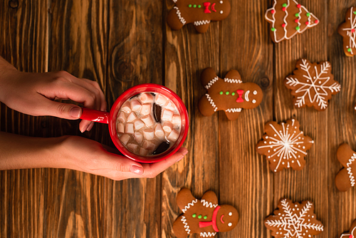 cropped view of woman holding cup of cocoa with marshmallows near gingerbread cookies on wooden surface