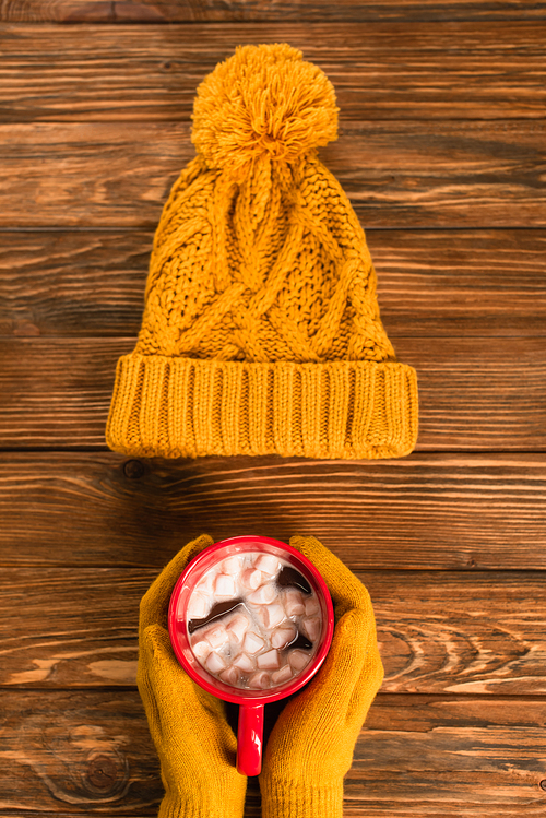 cropped view of person in yellow gloves holding cup of cocoa with marshmallows near hat on wooden surface
