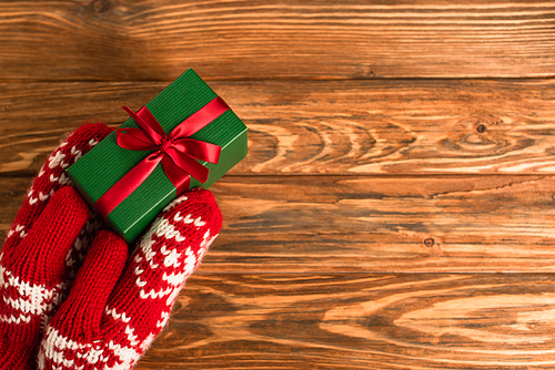cropped view of person in red mittens holding wrapped gift box above wooden surface
