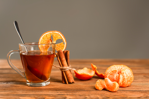 glass cup of tea with sliced orange near cinnamon sticks and peeled tangerine isolated on grey