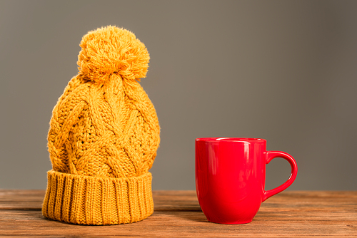 red cup of tea near knitted hat on wooden surface isolated on grey