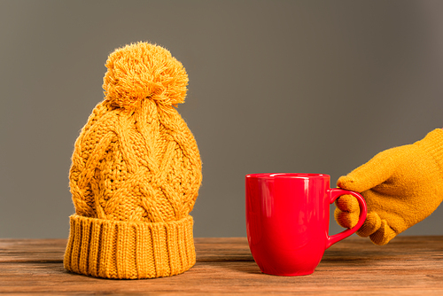 cropped view of person in glove holding cup of tea near knitted hat isolated on grey