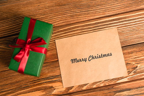 top view of greeting card with merry christmas lettering near wrapped green present on wooden surface
