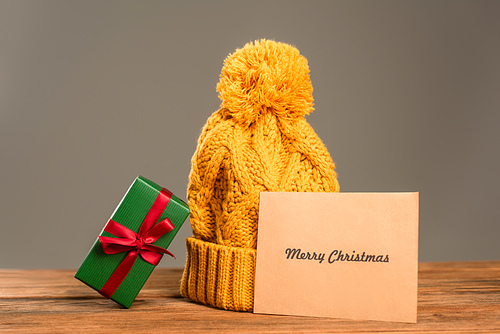 greeting card with merry christmas lettering near knitted hat and wrapped green present isolated on grey