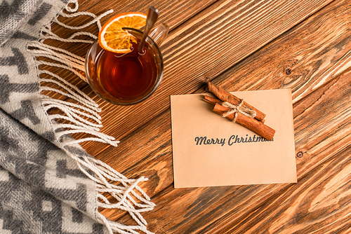 top view of cup of tea with sliced orange near cinnamon sticks on greeting card with merry christmas lettering on wooden surface