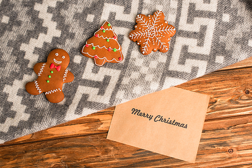 top view of gingerbread cookies on grey blanket with ornament near greeting card with merry christmas lettering on wooden surface