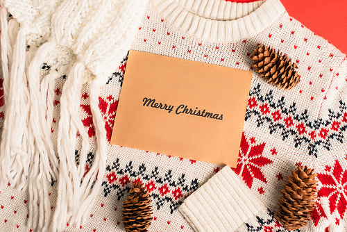 top view of greeting card with merry christmas lettering near pine cones on knitted sweater