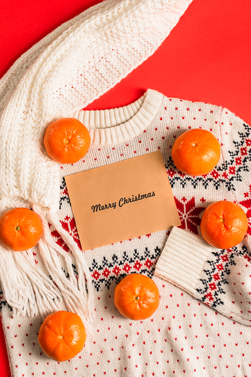 top view of greeting card with merry christmas lettering near tangerines on knitted sweater isolated on red