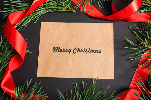 top view of greeting card with merry christmas lettering near fir branches and red ribbon on black