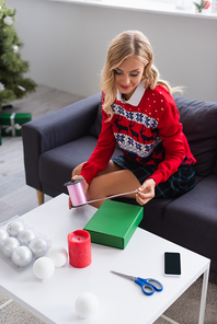high angle view of blonde woman with decorative ribbon near gift box and christmas balls on table