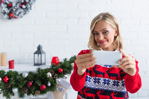 joyful woman in stylish sweater taking selfie on smartphone near blurred christmas garland