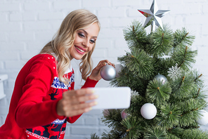 cheerful blonde woman in warm sweater taking selfie near christmas tree at home, blurred foreground