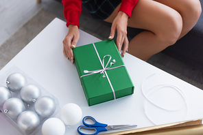 partial view of woman near green gift box, christmas balls and scissors on table