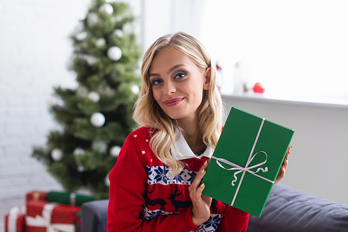 cheerful blonde woman in warm sweater holding green gift box near christmas tree on blurred background