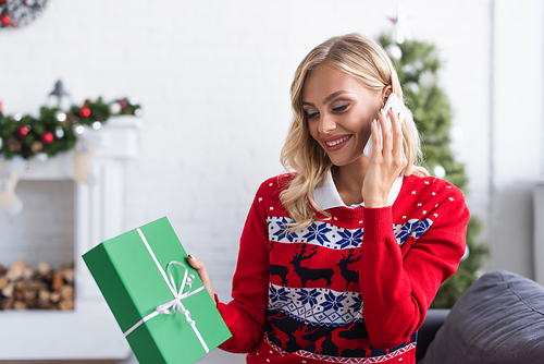 smiling woman in stylish sweater with ornament holding christmas present while talking on smartphone