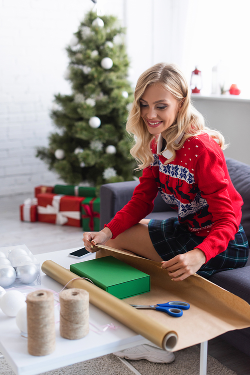 happy woman in warm sweater packing gift box into wrapping paper near presents under blurred christmas tree