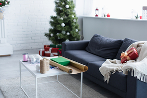 couch with warm blanket and gift box near table with twine, decorative ribbon and wrapping paper near blurred christmas tree