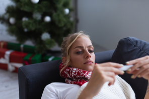 displeased and sick woman on sofa looking at thermometer near christmas tree on blurred background