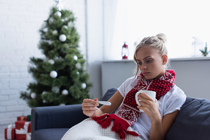 upset woman with warm drink and thermometer near christmas tree on blurred background