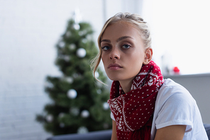 diseased woman in warm scarf looking at camera near blurred christmas tree