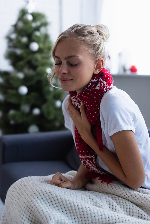 sick woman in warm scarf coughing on sofa with closed eyes near blurred christmas tree