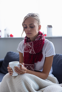 ill and sad woman in warm scarf sitting on sofa under blanket with cup of tea