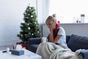 diseased woman in bad mood sitting with cup of warm drink near christmas tree on blurred background