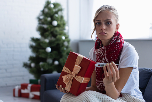 sad woman with gift box and thermometer  near christmas tree and presents on blurred background