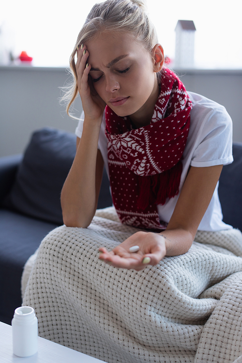 diseased woman in warm scarf sitting on sofa with closed eyes and pill in hand